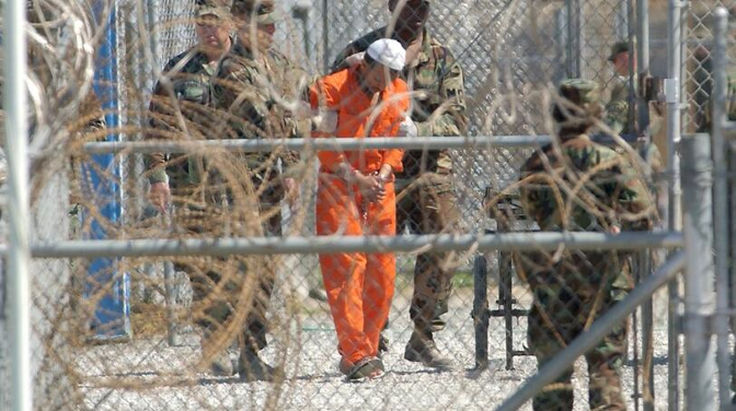 A man in orange jumsuit surrounded by guards
