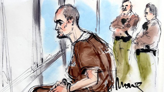 Two men standing behind man seated in brown jumpsuit