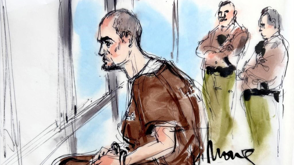 Two men standing behind man seated in brown jumpsuit